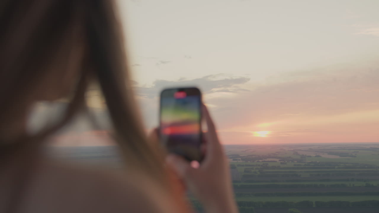 Close rear view of woman holding smartphone to capture glowing sunrise over vast green fields, soft golden light reflecting on device screen