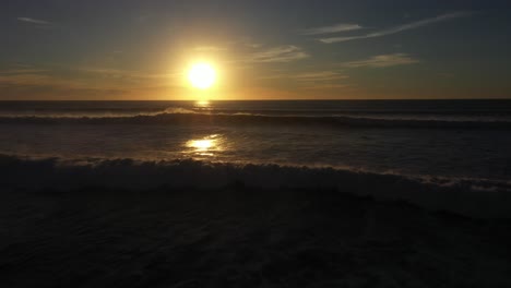 Breathtaking-drone-shot-captures-the-magical-moment-of-sunset-as-the-drone-flies-directly-over-the-crashing-waves,-with-the-mist-spraying-just-below-the-drone