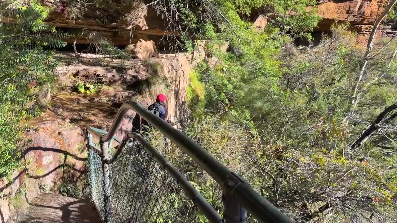 A woman with a backpack walks down a fenced mountain trail at Wentworth Falls, Australia.