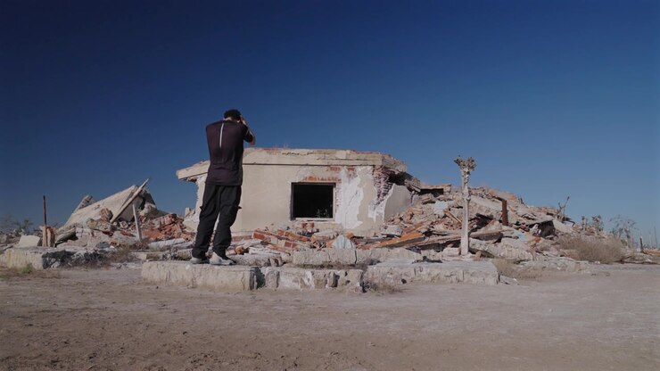 Un uomo sta in contemplazione davanti a un edificio crollato circondato da macerie e terra arida sotto un cielo blu vivido a Villa Epecuén, Buenos Aires, Argentina