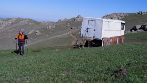 male backpacker walks past trailer bunkhouse toward camera in meadow