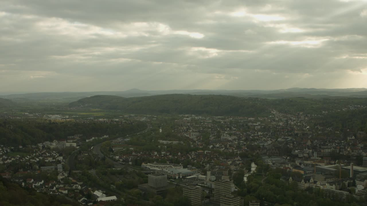 Overcast sky casts soft light over a sprawling European city nestled in lush hills, hinting at tranquility