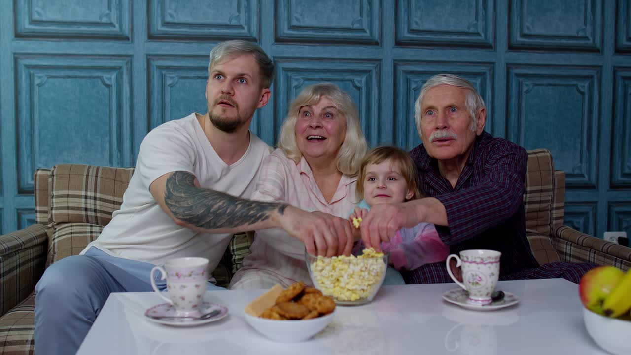Famiglia multigenerazionale guarda la TV con una bambina a casa, spaventata mentre guarda un film horror.