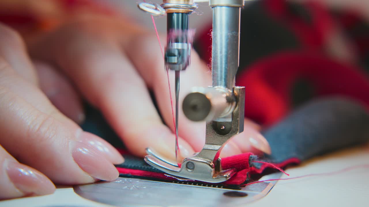 Primo piano di mani che guidano un tessuto rosso sotto l'ago della macchina da cucire. Dita ferme, concentrate, precise. Dettaglio della cucitura visibile, filo che attraversa il materiale