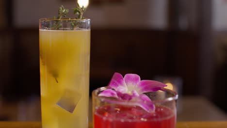 close up view of an orange cocktail with a thyme garnish and a red cocktail with a flower garnish in a japanese sushi restaurant with the video tilting up in slow motion