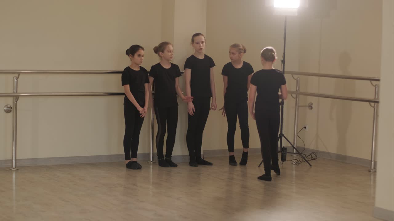 группа молодых студентов балета в черной танцевальной одежде практикует позиции в просторной балетной студии с деревянным полом и настенными решетками. сосредоточенные выражения и синхронизированные движения.