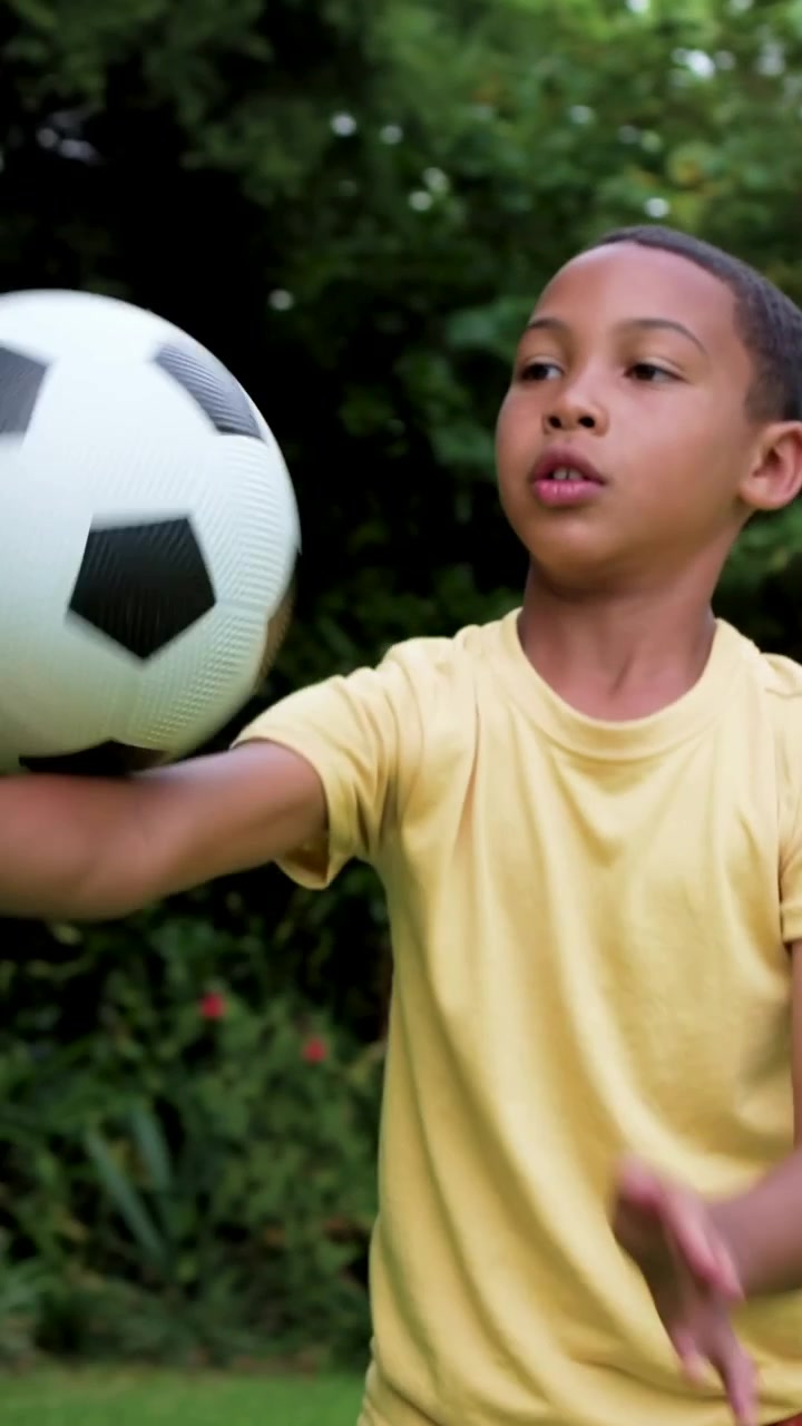 Vídeo vertical: Menino de camiseta amarela e shorts cor ferrugem jogando bola de futebol, pegando e acompanhando no quintal, treinando