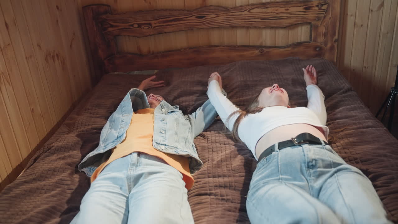 close up of two smiling ladies gazing warmly at each other in cozy wooden room filled with soft natural light, sharing joyful moment before falling back together on bed