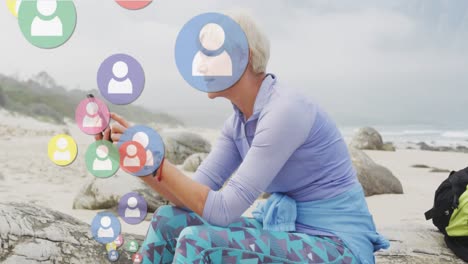 Animation-of-digital-people-icons-over-senior-woman-by-seaside-using-smartphone