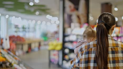 Rear-view-of-a-confident-brunette-girl-in-a-plaid-shirt-carries-her-small-child-in-her-arms-and-inspects-goods-in-a-supermarket-while-shopping