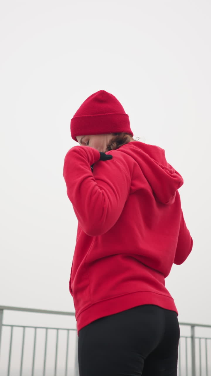 Back view of woman in red hoodie, black gloves, and red beanie turning arm and head to left in outdoor setting with iron railing and distant foggy atmosphere