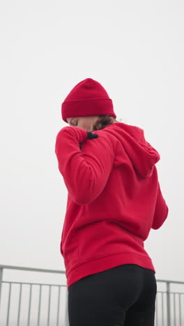 back view of woman in red hoodie, black gloves, and red beanie turning arm and head to left in outdoor setting with iron railing and distant foggy atmosphere