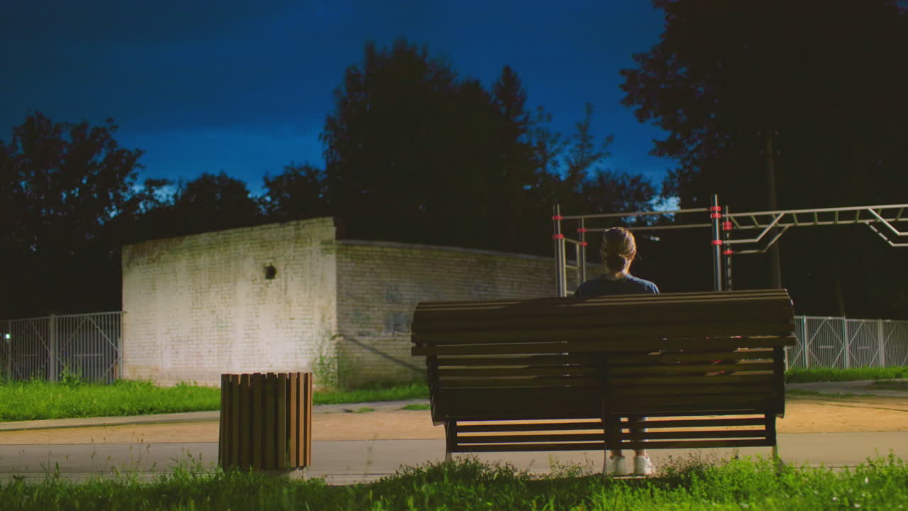 vista trasera de una mujer sentada al aire libre en un banco por la noche con la luz reflejándose suavemente en su cara, el fondo incluye una estructura de patio de recreo y una pared de ladrillo sombreada bajo un cielo azul profundo