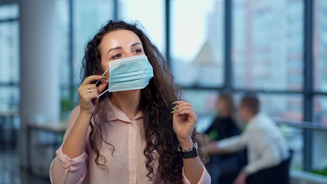 Dark-haired young woman is inside the office. Smiling lady puts on blue mask on her face and makes a gesture with her hands. Blurred background.