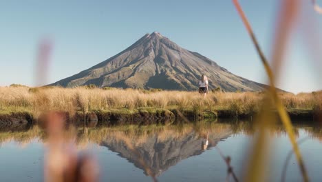 Blonde-female-walking-through-golden-grass-in-front-of-mirror-lake-with-mountain-in-background