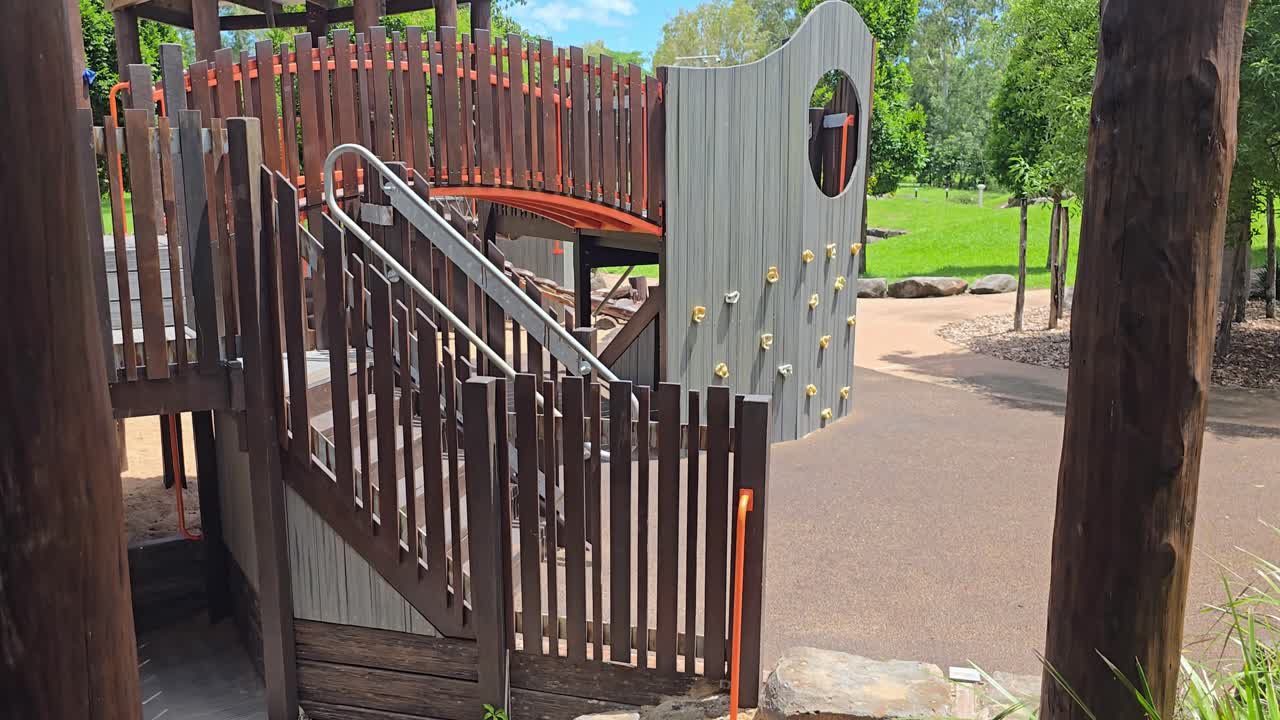 A children's wooden playground with a sand pit and rock climbing wall at Plantation Park in Ayr, North Queensland, with green grass and blue sky in the background