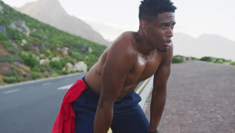 hombre afroamericano haciendo ejercicio al aire libre apoyándose en sus rodillas en una carretera costera