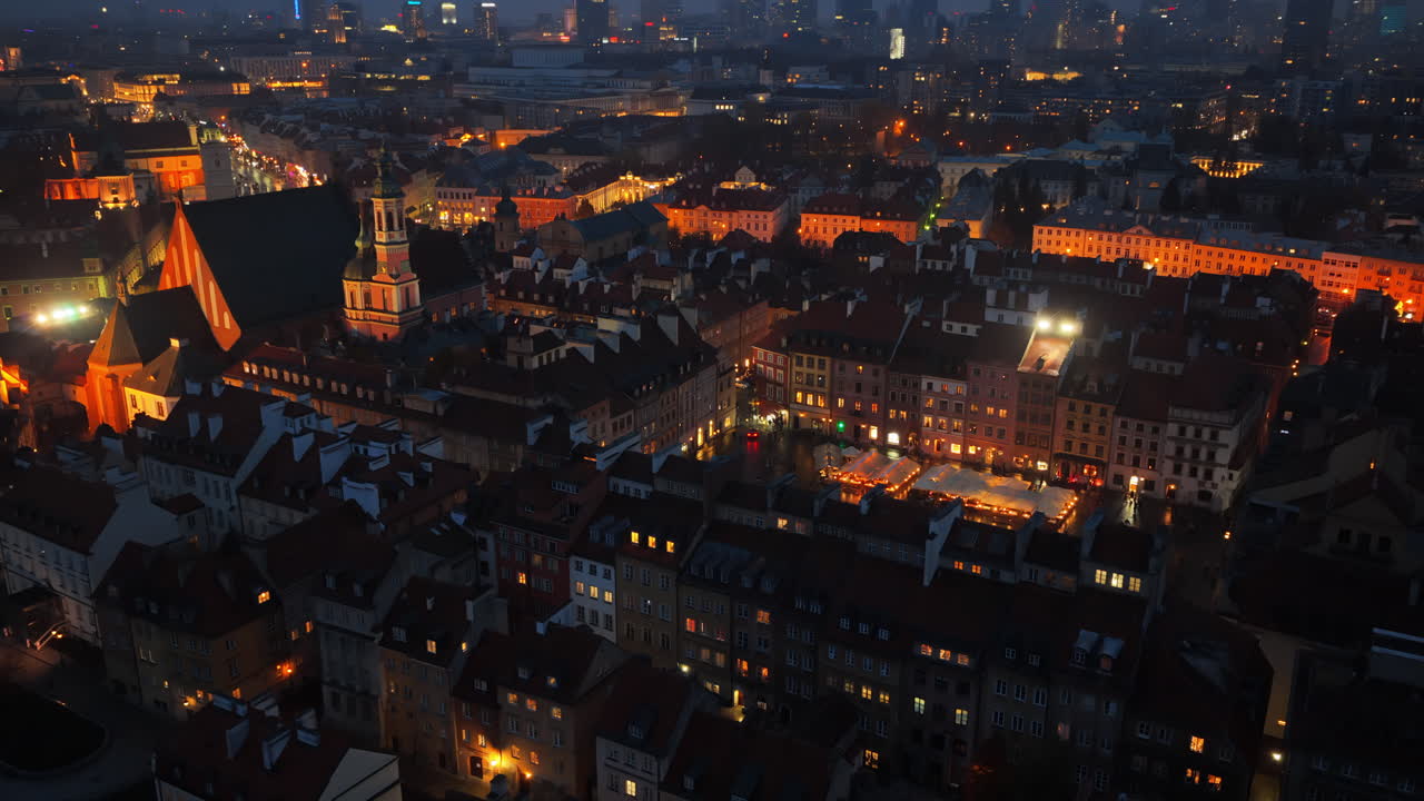 Aerial drone view of Warsaw's Old Town Market Square, showing illuminated vendor stalls, people walking, and the geometrical layout of the historic square