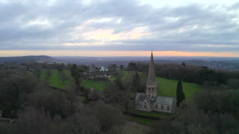 una toma aérea volando más allá de una antigua iglesia inglesa en una colina en ranmore common, en dorking al amanecer