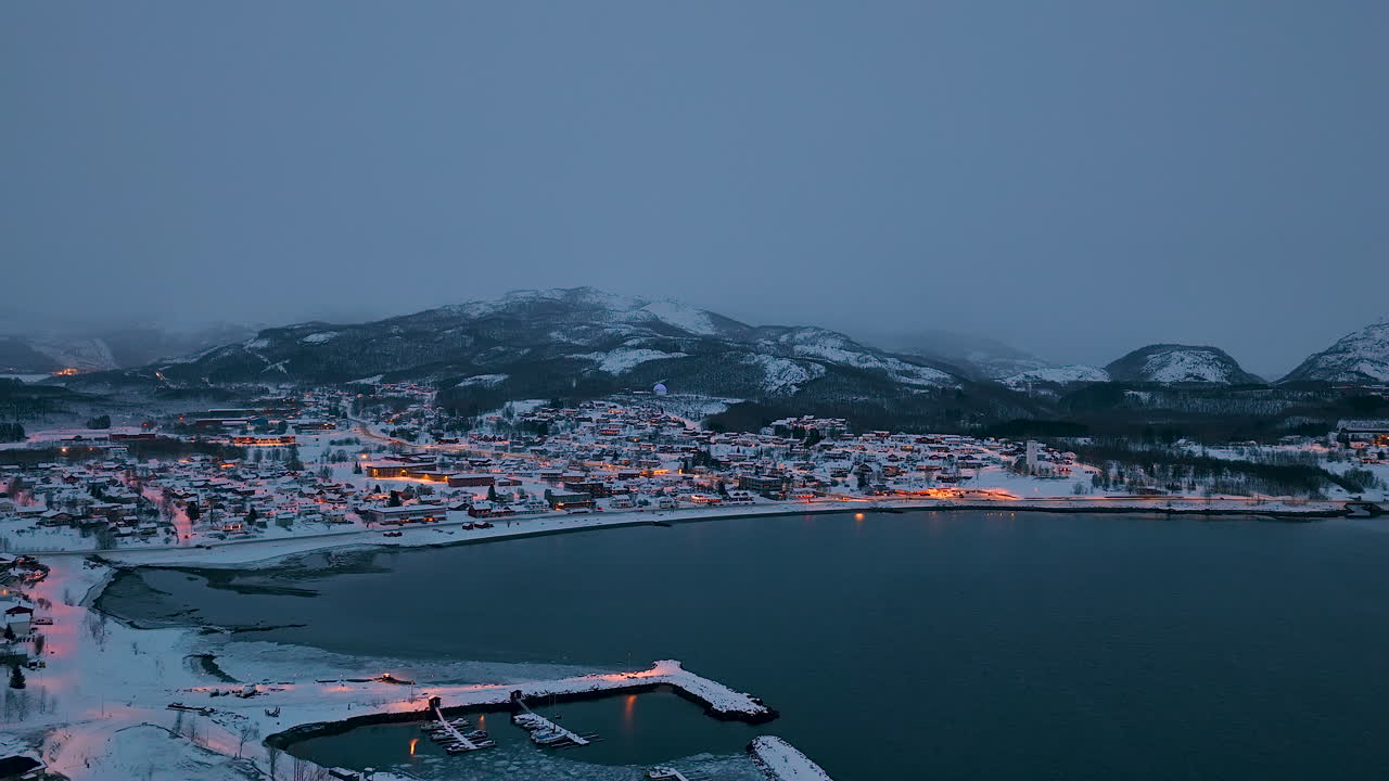 Fantastisk flyfoto av en snødekt norsk kystlandsby i skumringen. Scenen viser glødende bylys mot et mørkeblått hav og tåkete fjell i Lofotarkipelet.