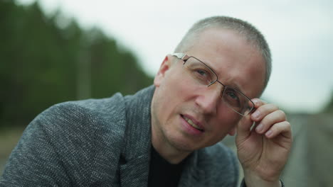 a close-up view of a middle-aged man adjusting his glasses while looking into the camera, he is outdoors, wearing a gray jacket, and appears thoughtful and focused