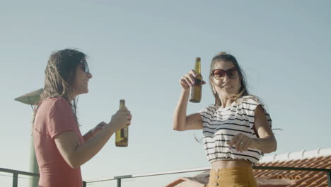 niñas caucásicas felices sosteniendo cervezas y bailando en la azotea durante una fiesta al aire libre