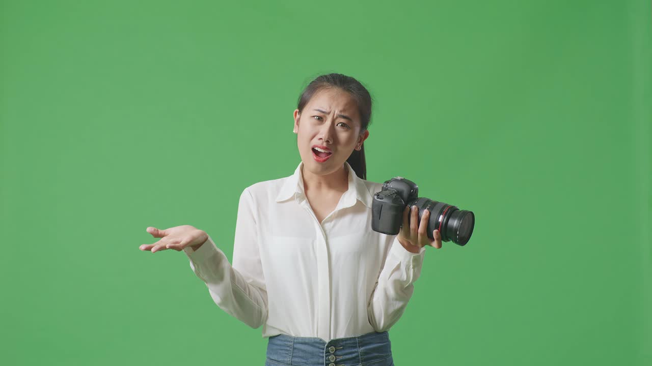혼란스러운 아시아 사진작가는 손에 카메라를 들고, 왜라고 말하고 스튜디오에서 녹색 화면 배경에 서서 의심스럽게 몸짓을 합니다.