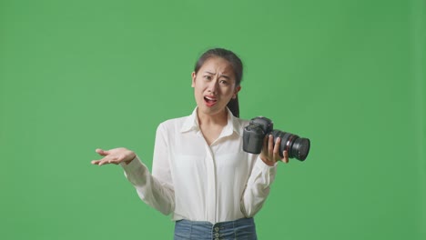 혼란스러운 아시아 사진작가는 손에 카메라를 들고, 왜라고 말하고 스튜디오에서 녹색 화면 배경에 서서 의심스럽게 몸짓을 합니다.