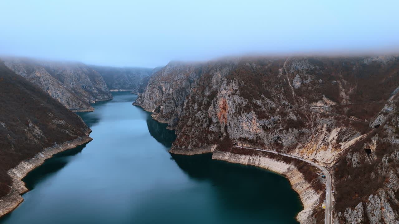 Strada tortuosa lungo un canyon fluviale con scogliere rocciose e nebbia. Ripresa aerea di una strada di montagna che costeggia un canyon fluviale turchese sotto uno spesso strato di nuvole e nebbia