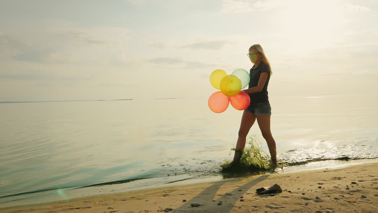 una joven despreocupada camina por la playa en el hermoso cielo sostiene globos en cámara lenta