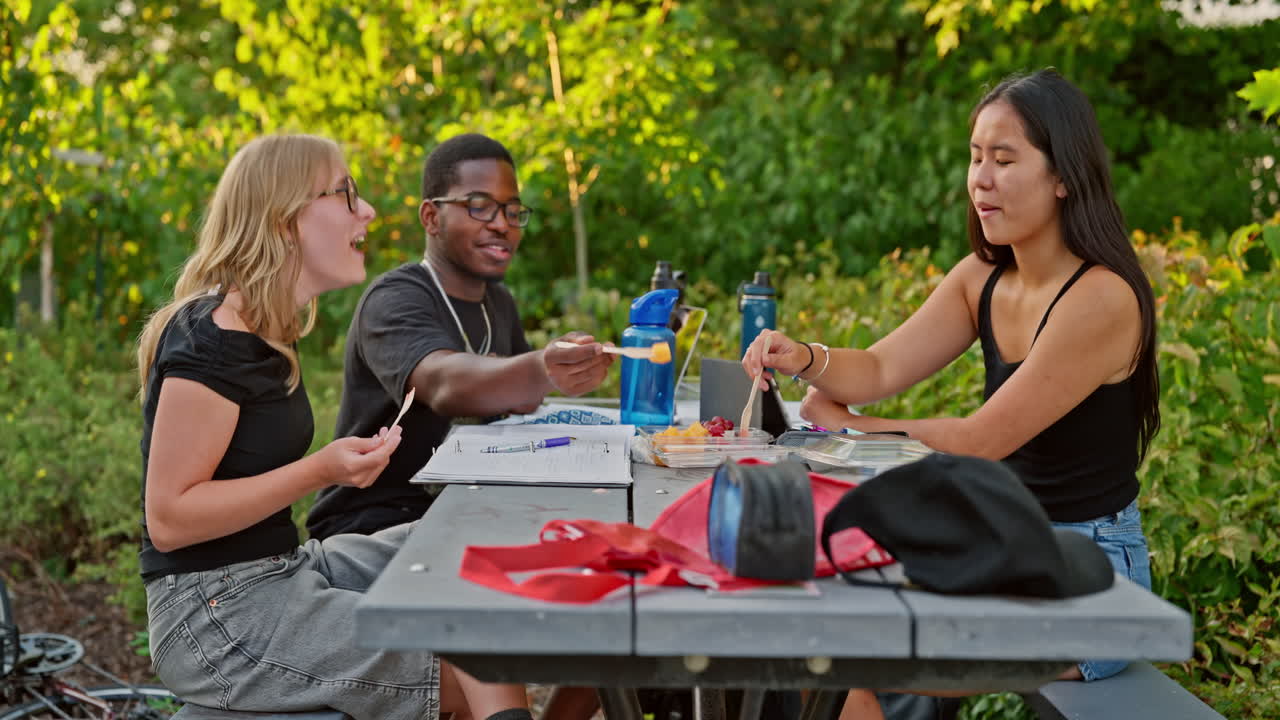 Three expressive Gen Z college students eating fresh fruits and interacting with joy at a park table. Positive emotions, teamwork and friendship under natural light in a relaxed learning environment