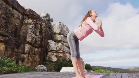 caucasian woman practicing yoga, stretching and touching floor, standing on deck on mountainside