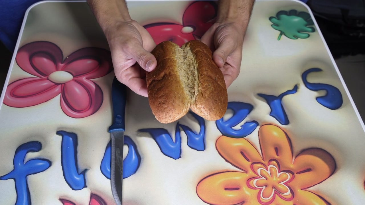 Top down close up view of male hands using a knife to slice open a fresh brown wheat bread roll. Preparing a simple meal indoors over a bright colorful floral kitchen table during daytime.