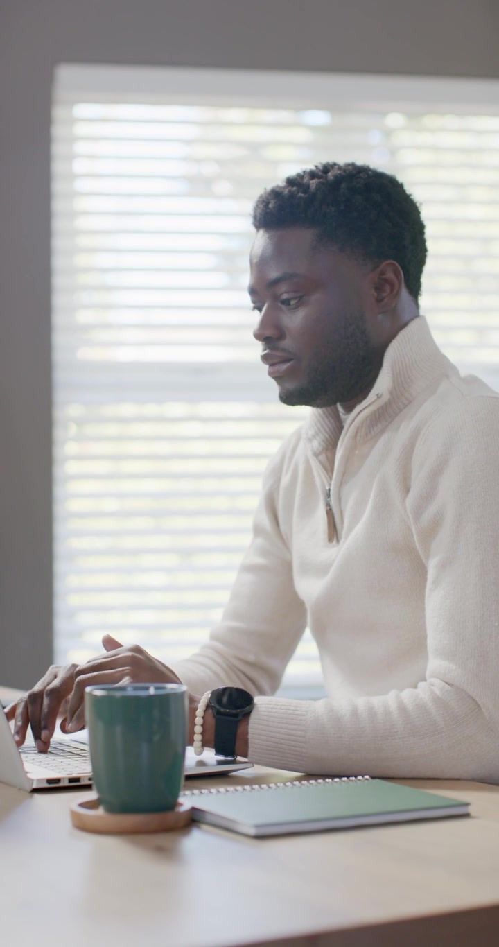 Vertical video: Man stretching, reaching laptop and mug, typing and starting work at desk by window