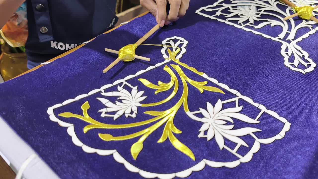 Seorang perajin terampil menampilkan sulaman Tekat Melayu tradisional. Ia dengan cermat menyulam benang emas di atas kain beludru mewah. Kerajinan kuno yang langka ini mewakili bentuk seni warisan yang semakin menghilang.