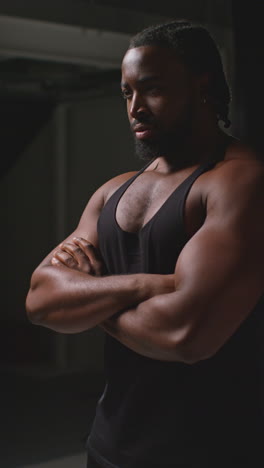 Retrato-En-Video-Vertical-De-Un-Atleta-Masculino-Con-Chaleco-Deportivo-Y-Físico-Musculoso,-Con-Los-Brazos-Cruzados-Entrenando-Para-Un-Evento-Deportivo-Contra-Un-Fondo-De-Estudio-Negro