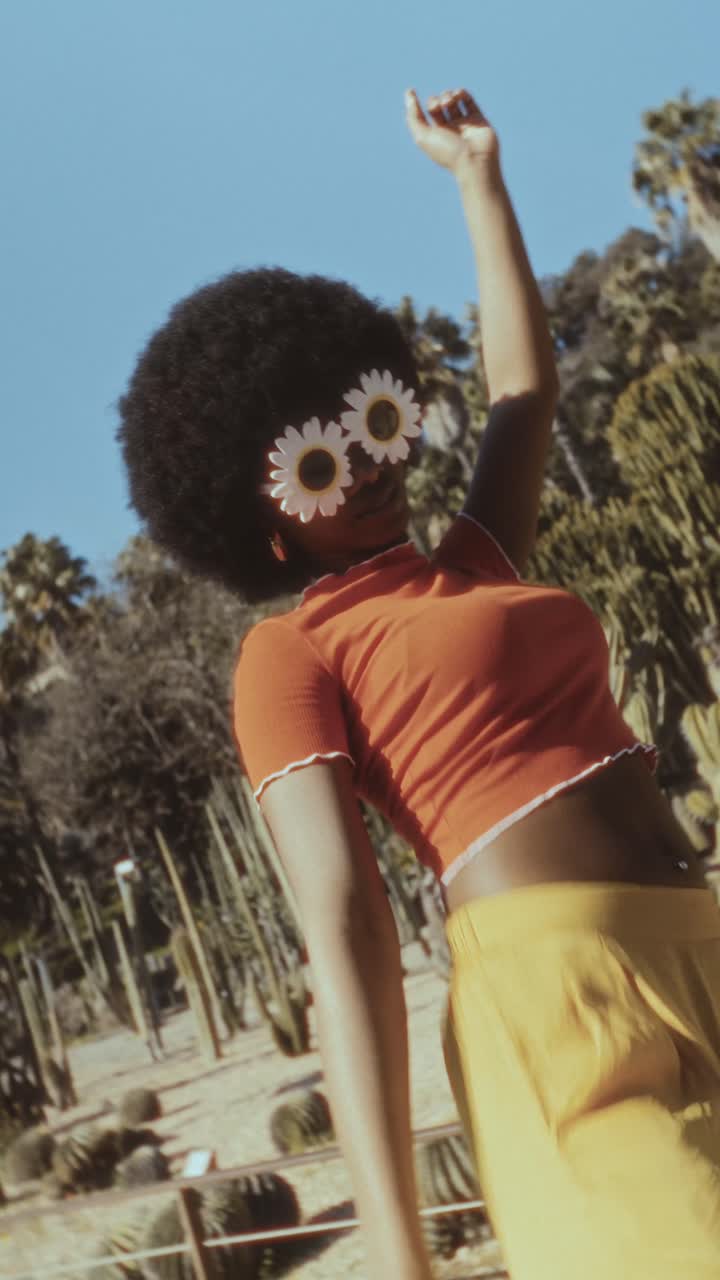 Giovane donna giocosa con afro e occhiali da sole a margherita che balla all'aperto