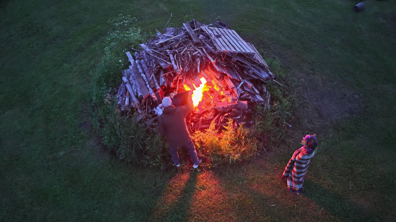 People light traditional Līgo bonfire using wooden pallets and branches on grassy field