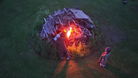 people light traditional līgo bonfire using wooden pallets and branches on grassy field