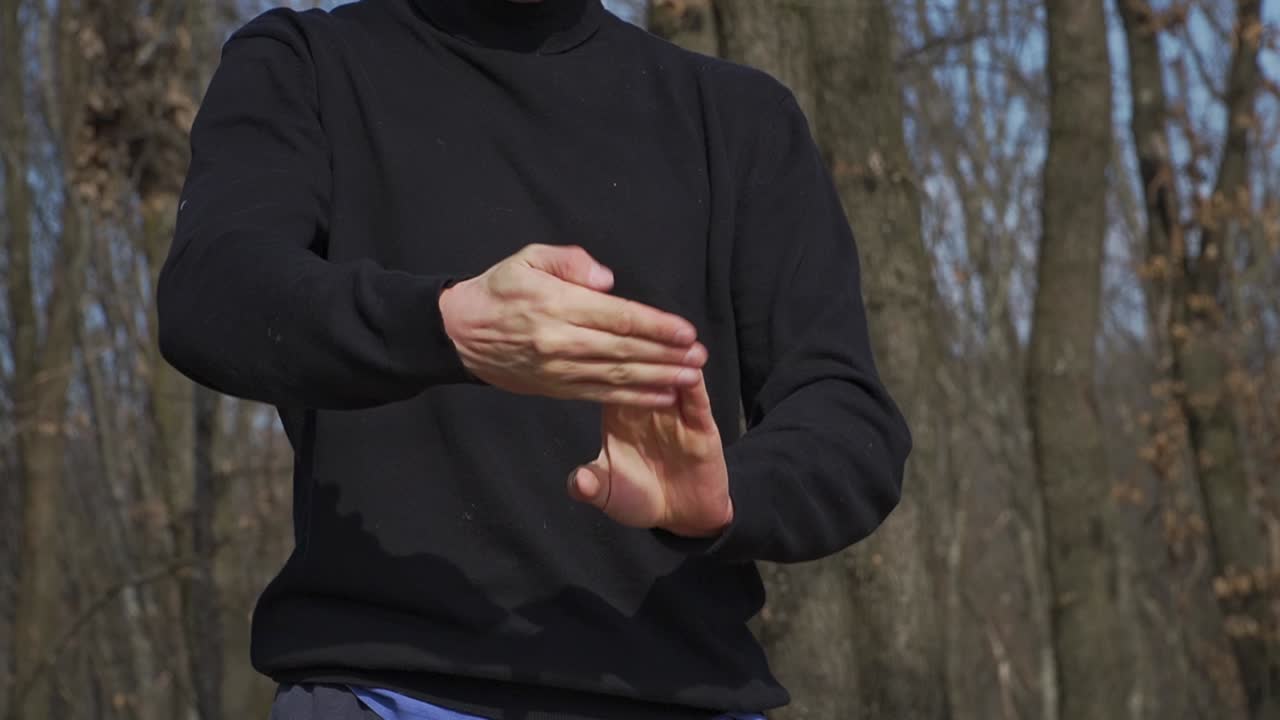 A martial artist in a black outfit stretches his arms, wrists, sholders and fingers, set against a forest backdrop. A gesture of discipline, strategy, and inner strength before battle
