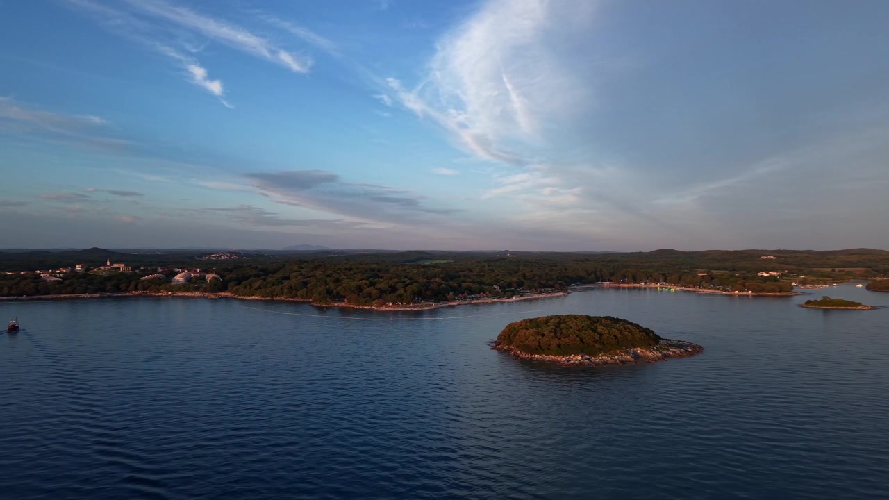 Piccola isola boscosa galleggia su calme acque adriatiche accanto alla costa istriana coperta di pini al tramonto dorato, con la scia di una barca e nuvole sottili vicino a Orsera, Istria, Croazia. Ampia inquadratura aerea con drone.