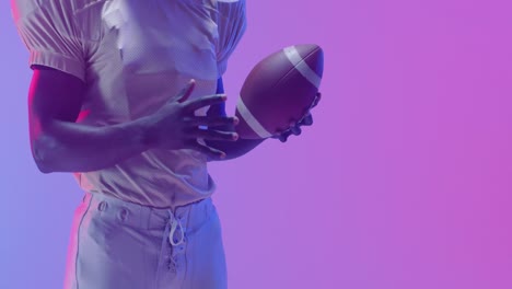 Video-of-midsection-of-african-american-american-football-player-with-ball-over-purple-background