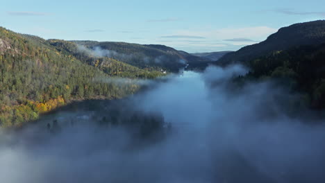 aerial drone view over a lake and waterfall, in middle of hills and foliage forest, foggy, autumn morning, in south norway