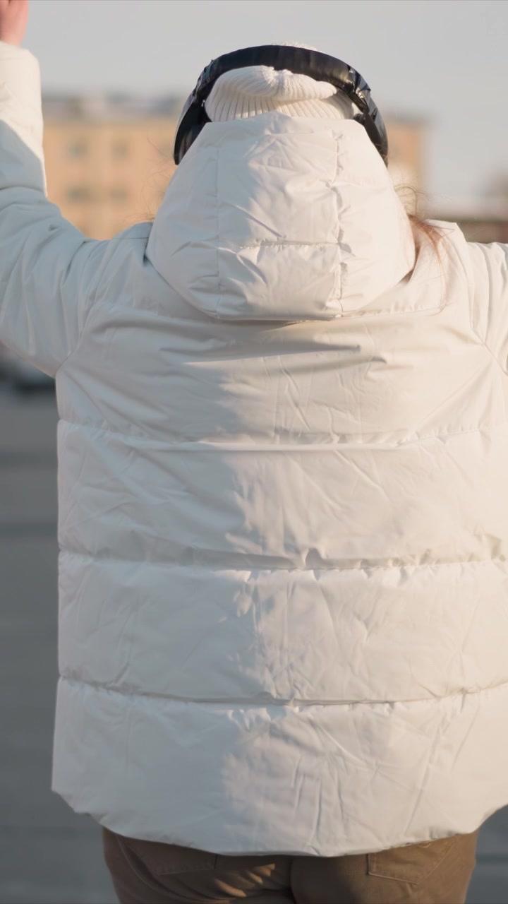 Woman in white puffer jacket and headphones stands outdoors raising her arms to signal vehicles. background shows urban setting with buildings and cars visible under soft daylight lighting