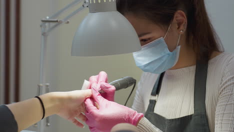 nail technician wearing mask, white shirt, and apron carefully using electric tool to shape customer's nails under focused lamp light during professional manicure session in clean salon environment
