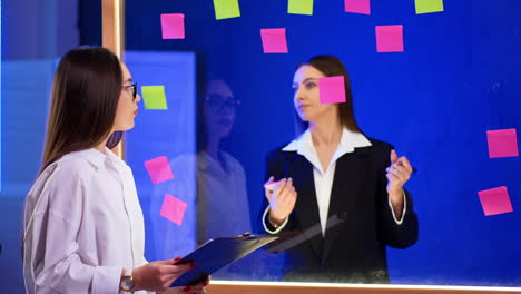 brunette holding a clipboard stands looking at glass wall. woman behind the board puts stickers on it. brainstorming in the office.