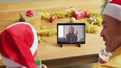 padre y hijo caucásicos con sombreros de santa usando una tableta para una videollamada de navidad con una mujer en la pantalla