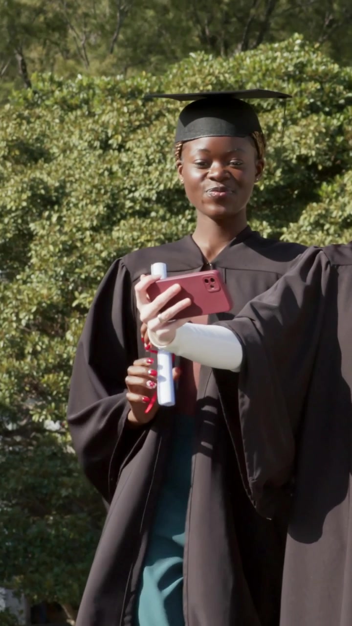 Vertical video: Walking grad in gown cap framing selfie on campus, holding pink phone and diploma