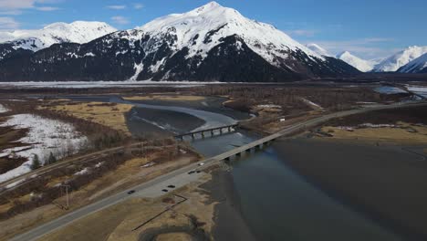 4K-Drone-slowly-panning-up-over-a-bridge-revealing-a-tall-whitecapped-mountain-in-Alaska-early-spring-with-blue-skies-,-reveal-shot-and-establishing-shot
