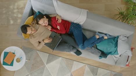 young couple is lying down on a couch, using a laptop. they are happy and smile. cozy living room with modern interior with plants, table and wooden floor. top view.